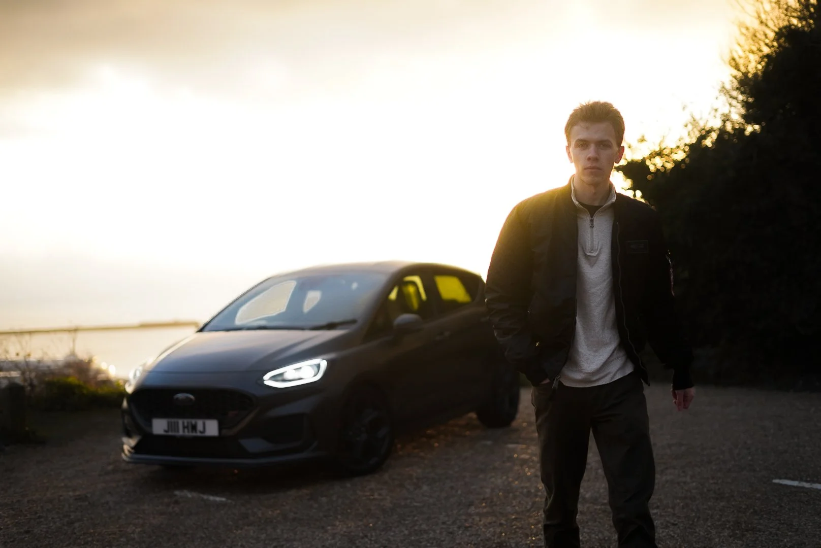 A young man stands outdoors near a parked black car during sunset, with a body of water and trees in the background.