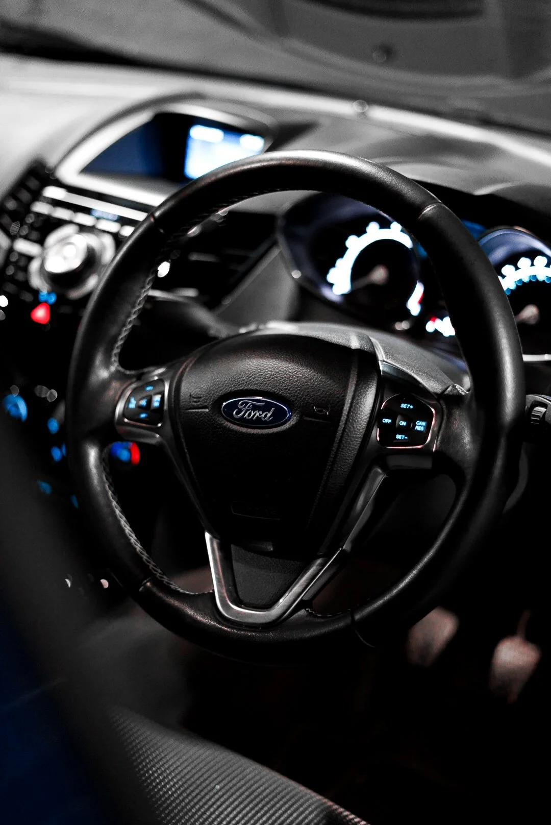 Close-up of a steering wheel with the Ford logo inside a car dashboard, illuminated instrument panel in the background.