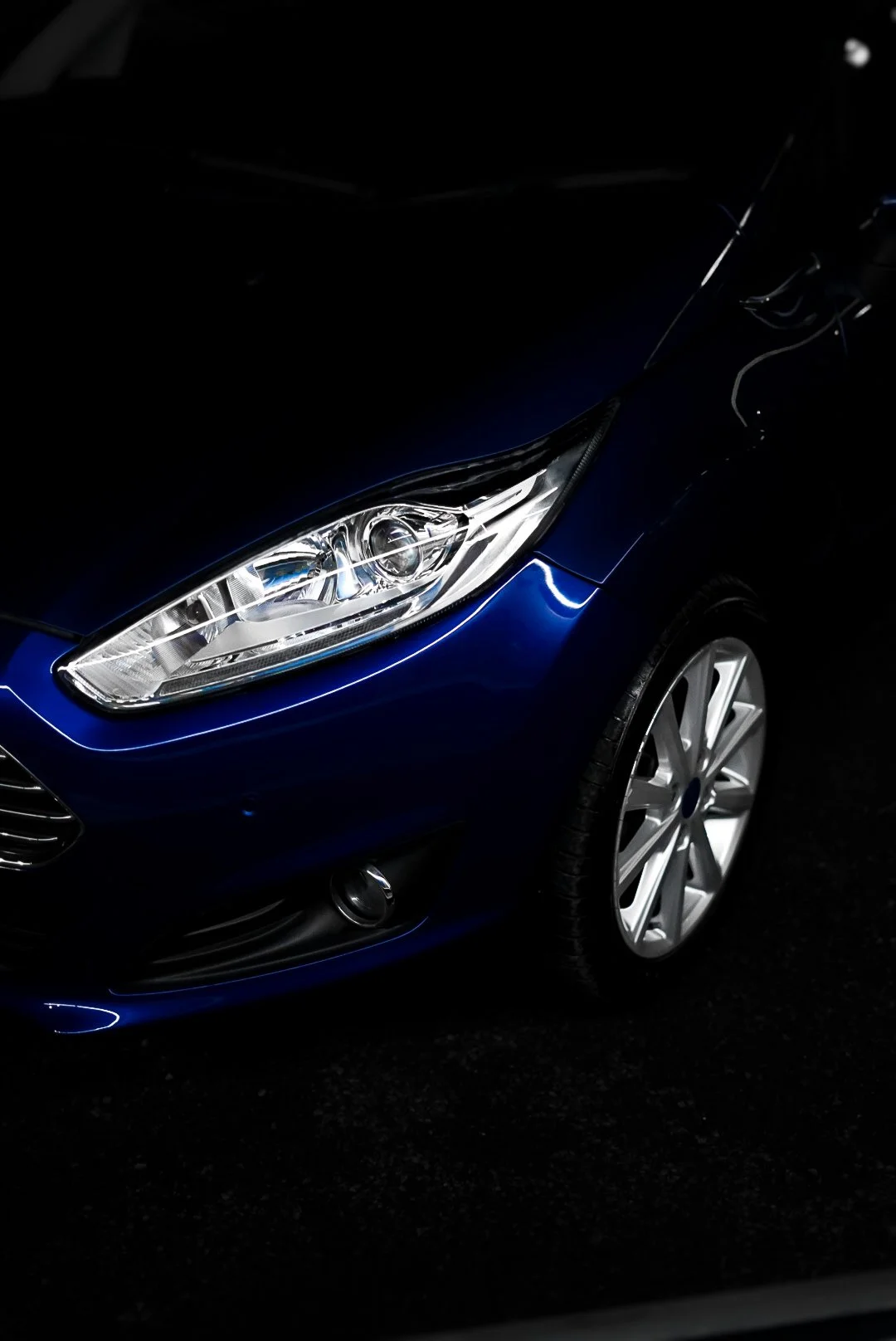 Close-up of front right corner of a dark blue car, showing headlight, wheel, and part of bumper, in a dark setting.