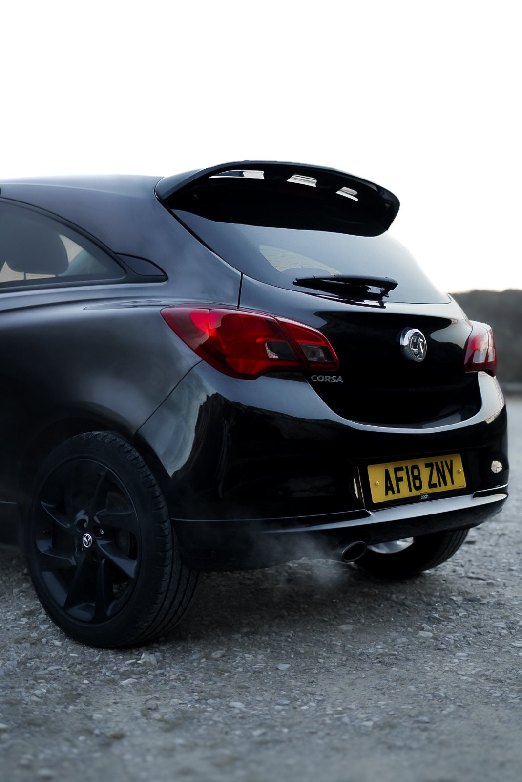 Rear view of a black Vauxhall Corsa with a yellow license plate, parked on gravel, with a sunset or sunrise sky in the background.