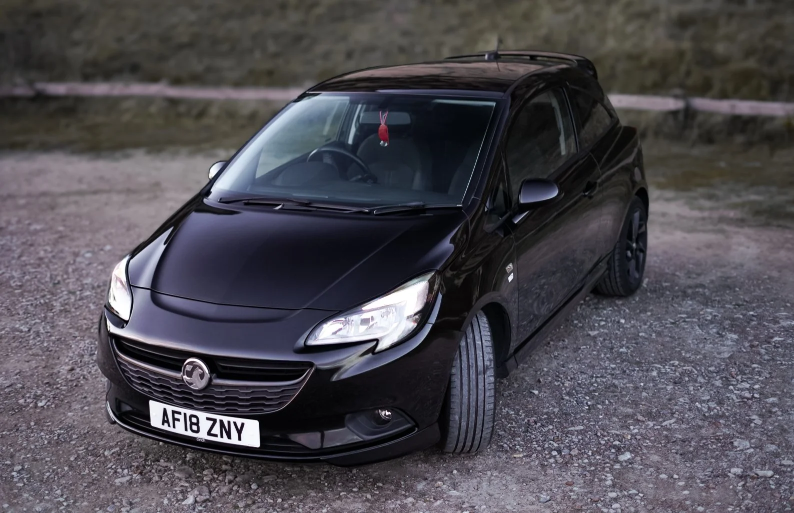 A black hatchback car parked on a gravel surface, with a grassy hill in the background.