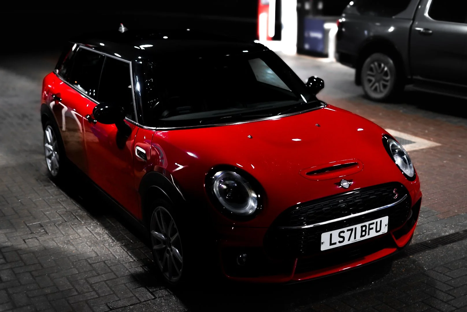 Red Mini Cooper parked on a brick pavement at night with other cars and vending machines in the background.