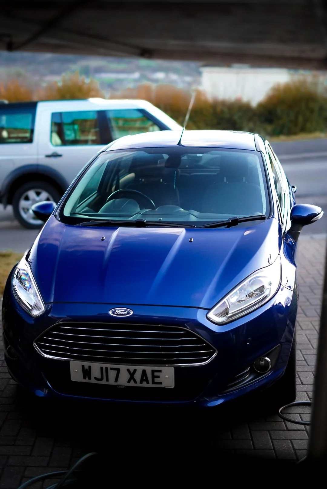 Blue Ford car parked outdoors under a bridge, with a silver SUV visible in the background.