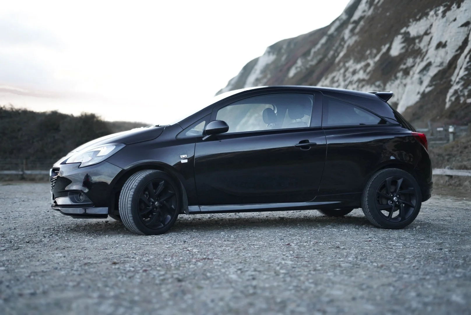 A black compact hatchback car parked on a gravel surface with mountains and cloudy sky in the background.