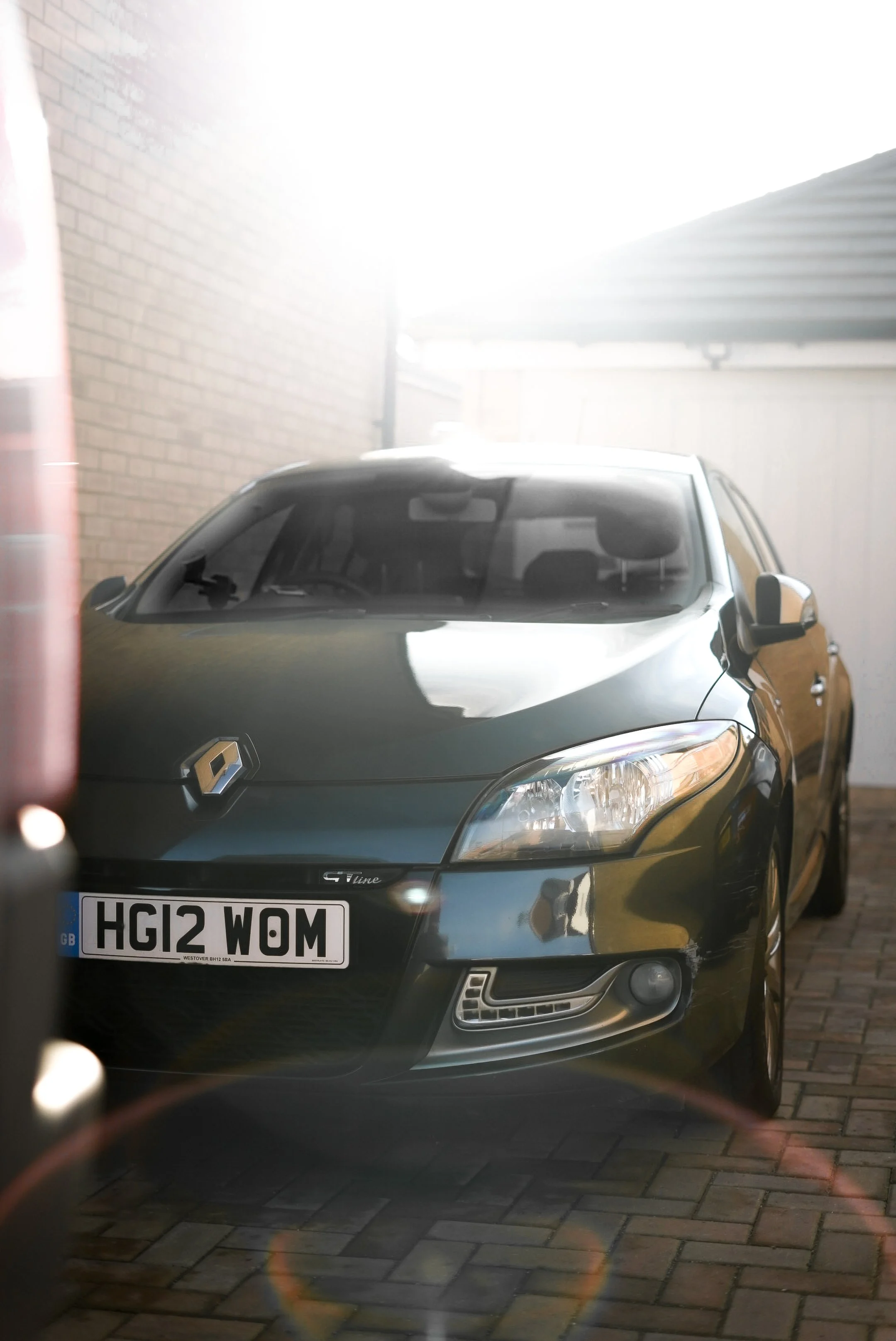 Black Renault car parked on a brick driveway next to a white garage and a brick wall, with sunlight creating a lens flare.