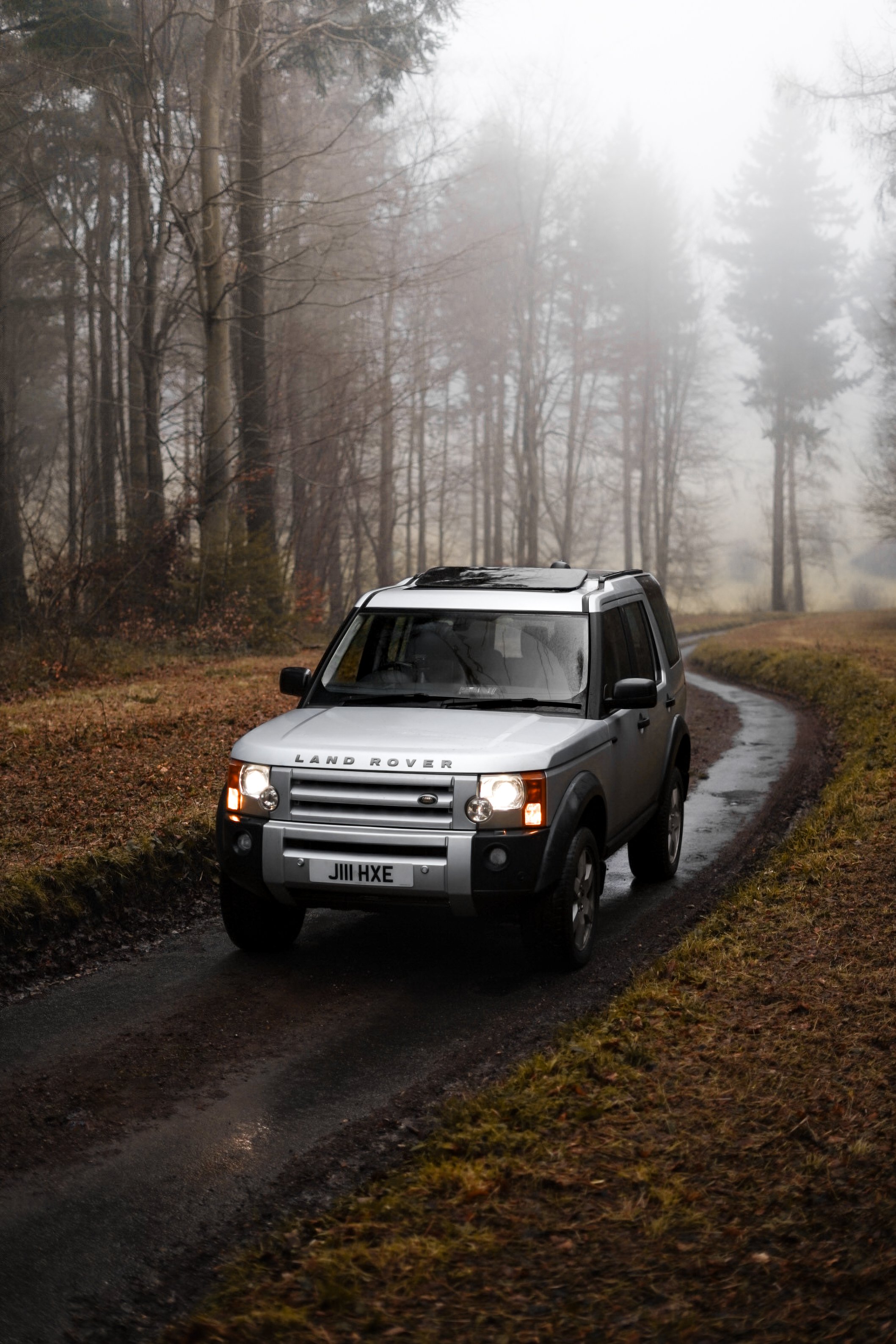 A silver Land Rover driving on a narrow muddy path through a foggy forest with leafless trees and brown grass.