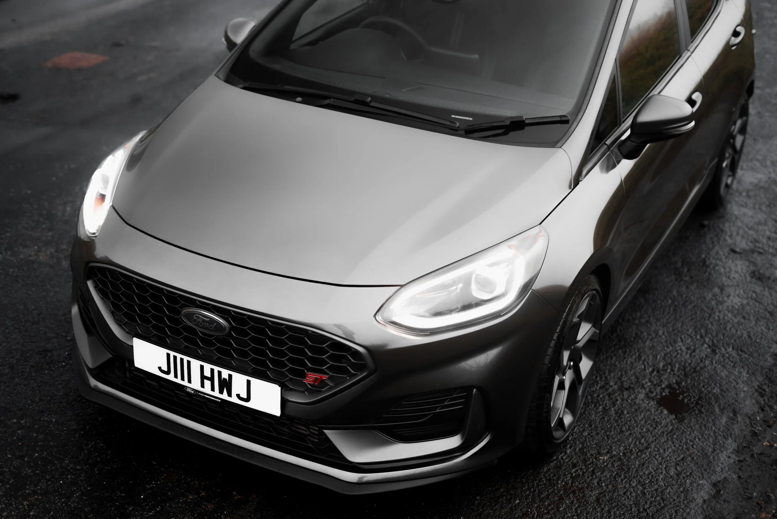 A silver Ford hatchback car parked on a wet asphalt surface, viewed from above at an angle.