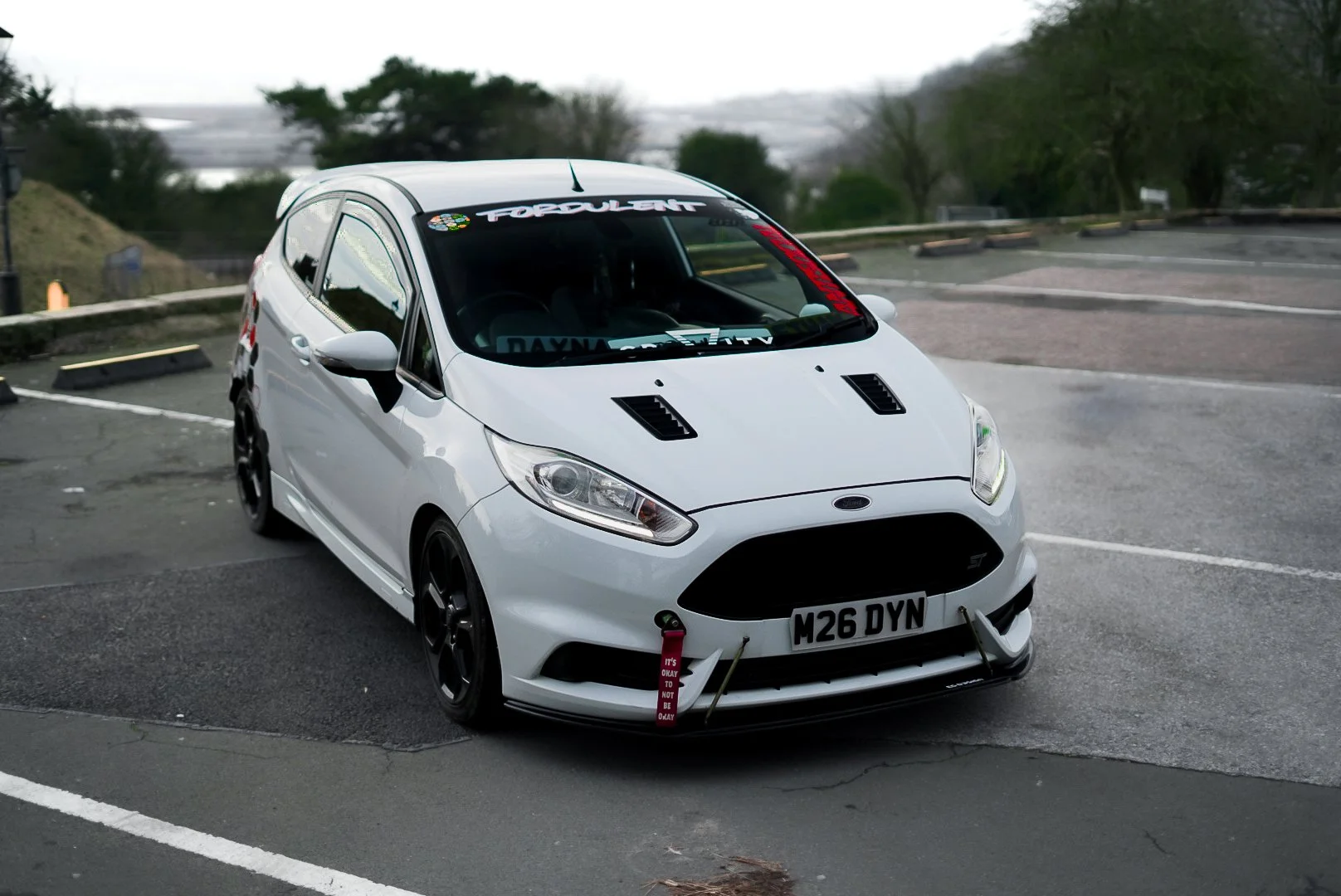 White modified Ford Fiesta parked in a parking lot with custom front bumper, hood vents, and racing decals, including a pink tag hanging from the front and a blacked-out grille.
