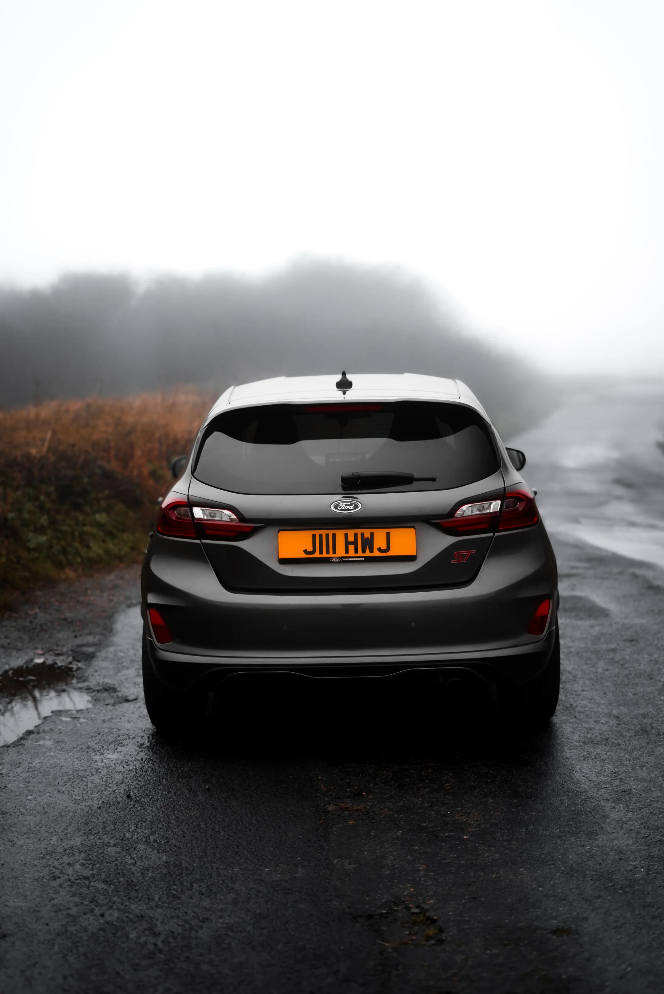 Black Ford hatchback car on a foggy road with wet pavement and puddles, surrounded by foggy landscape and trees in the distance.