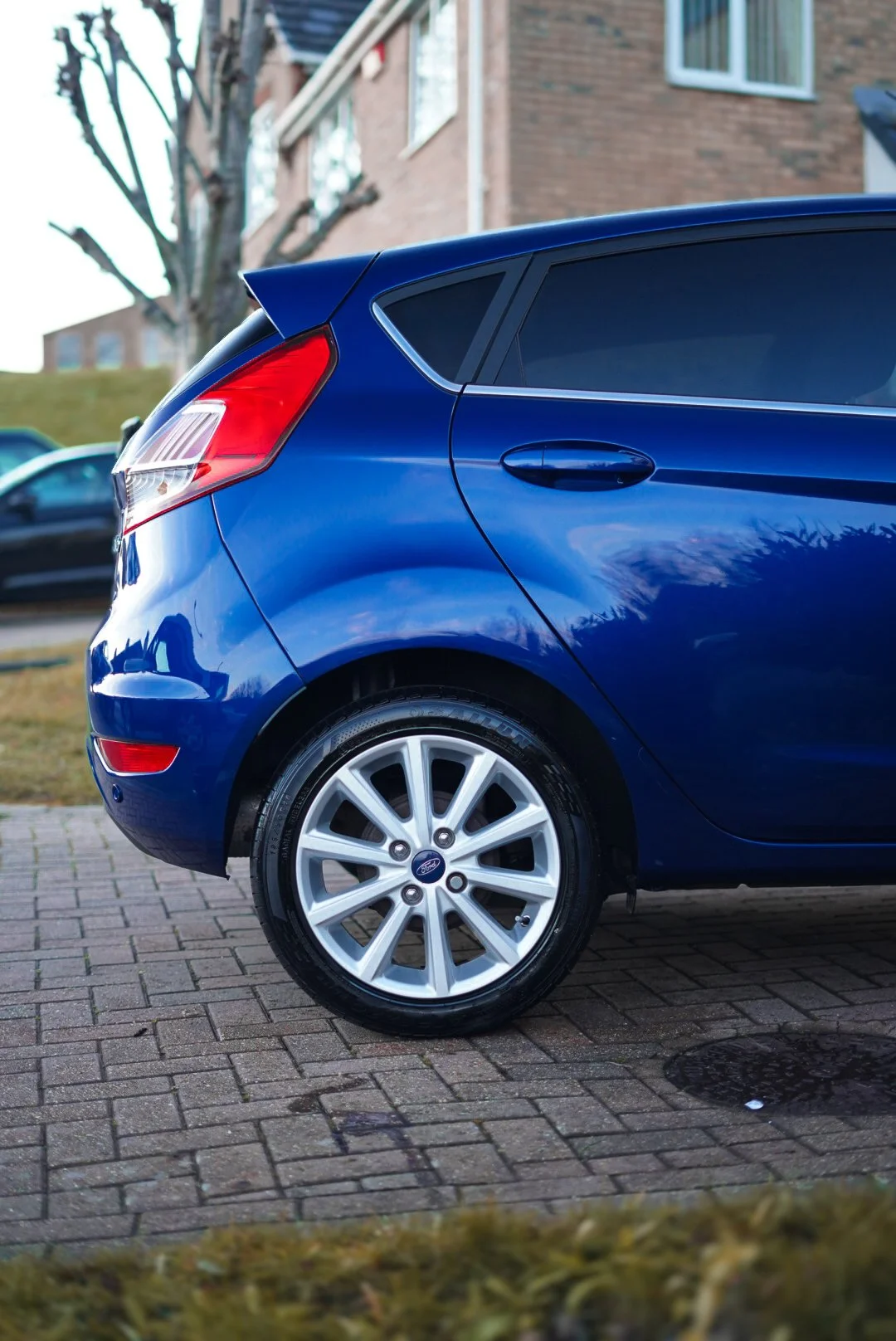 A blue hatchback car parked on a brick driveway, with a tree and residential buildings in the background.