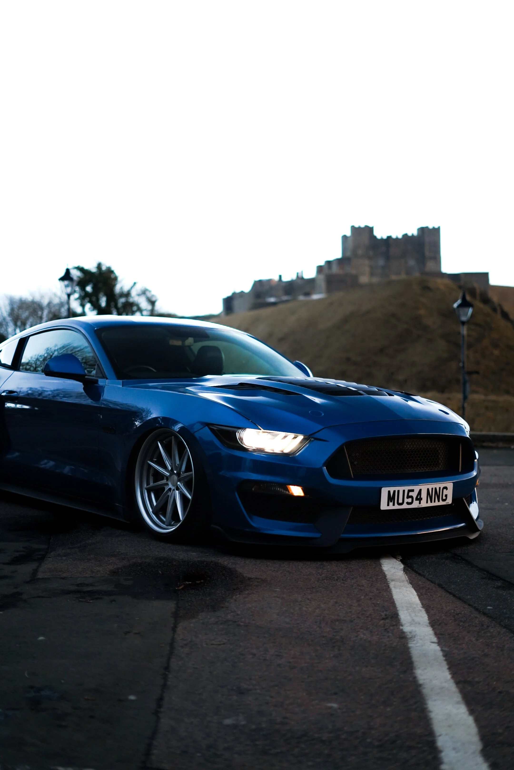 A blue sports car parked on a street with a castle on a hill in the background, early evening or dusk lighting.