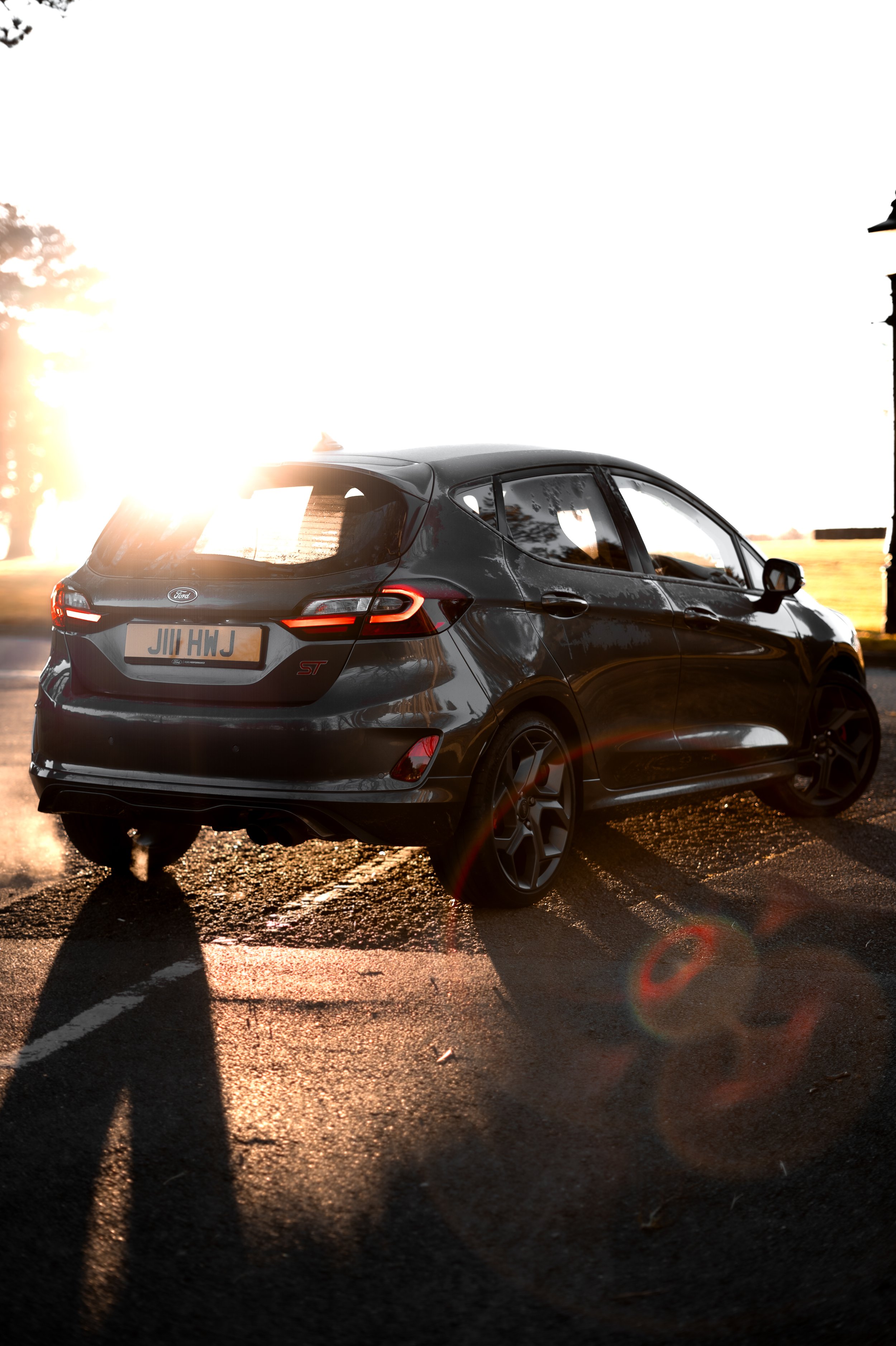 A black Ford Fiesta ST car parked in a parking lot during sunset, with sunlight creating a lens flare effect.