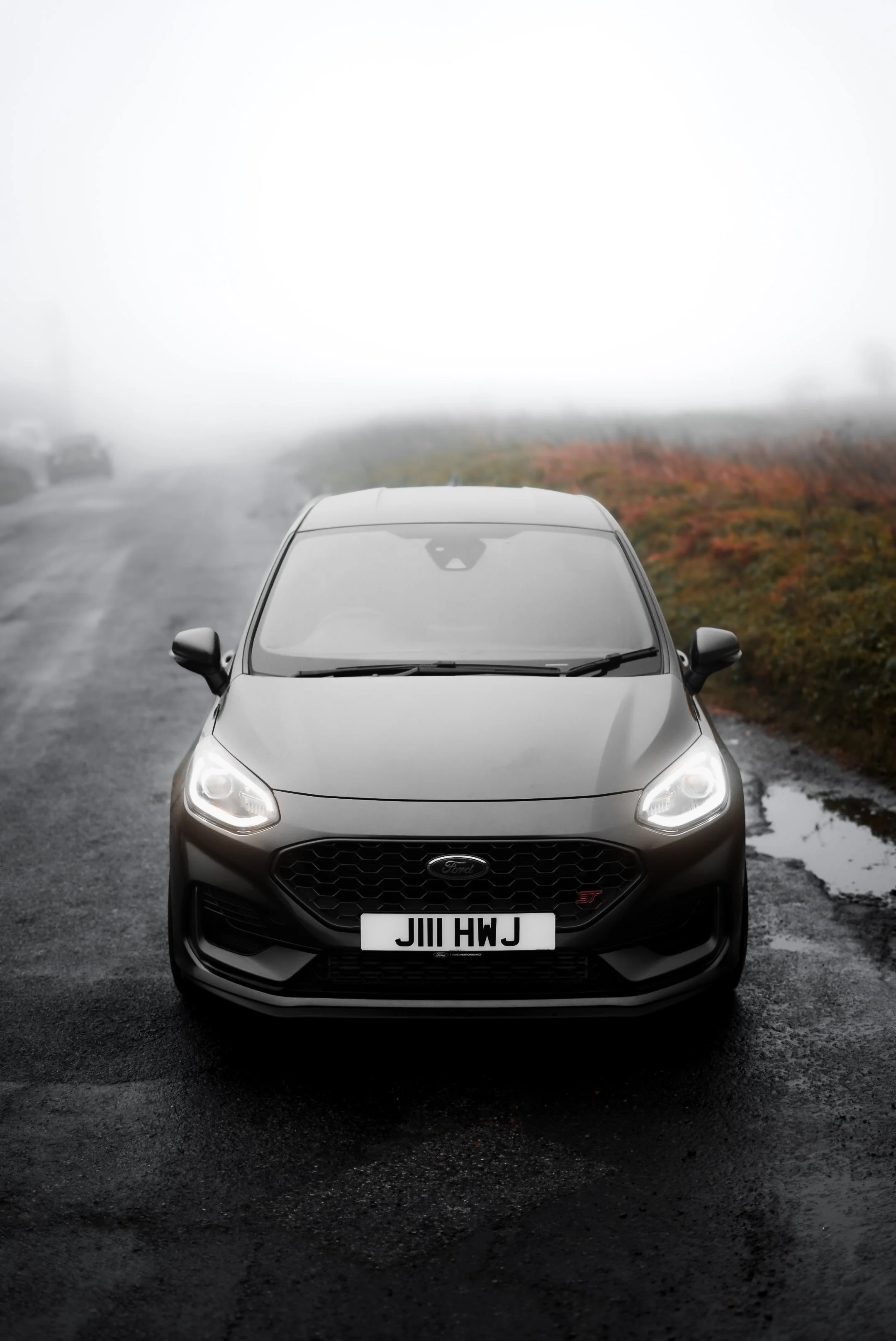Front view of a black Ford car with a license plate JIII HWJ on a wet, foggy road.