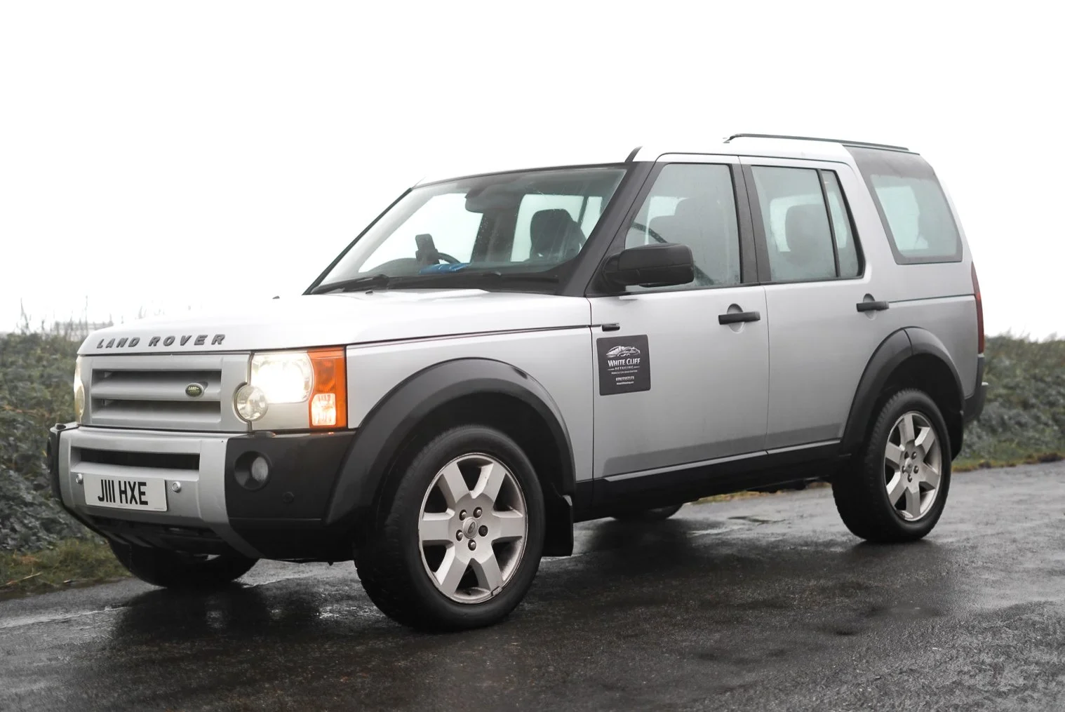 A silver Land Rover Freelander parked on a wet road with overcast skies in the background.