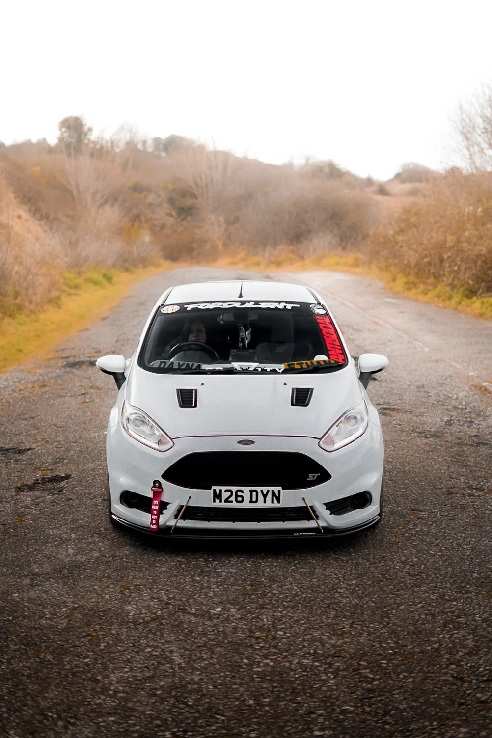 A white Ford Fiesta ST race car with a custom front license plate M26 DYN parked on a rural gravel road with leafless trees and hills in the background.