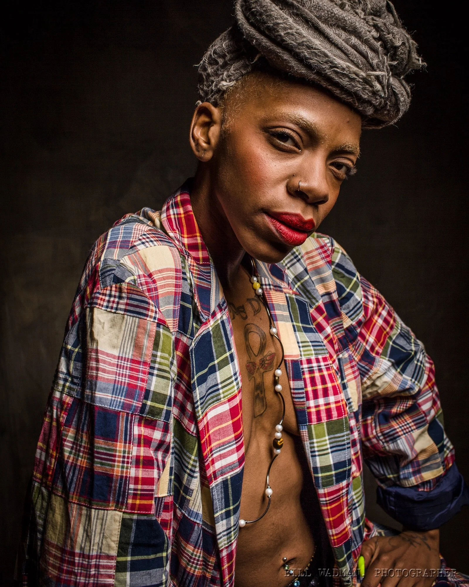 Portrait of a confident woman with gray dreadlocks, wearing a colorful plaid shirt unbuttoned to reveal tattoos and wearing a necklace, with a dark background.