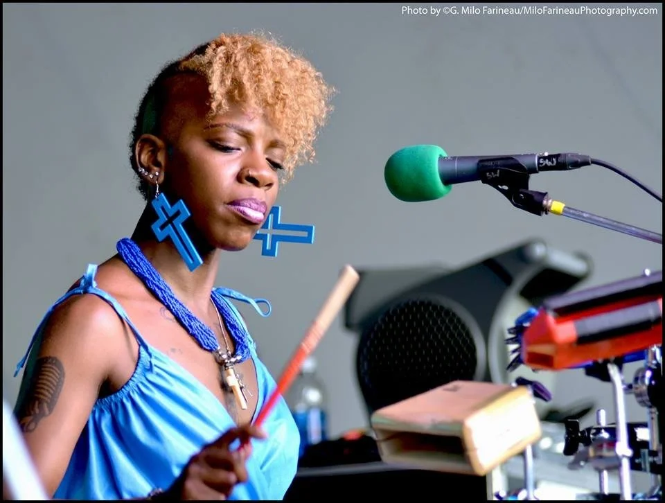 Cole Williams with short, curly blond hair, blue jewelry, and earrings shaped like crosses, playing bongos with a green microphone nearby.