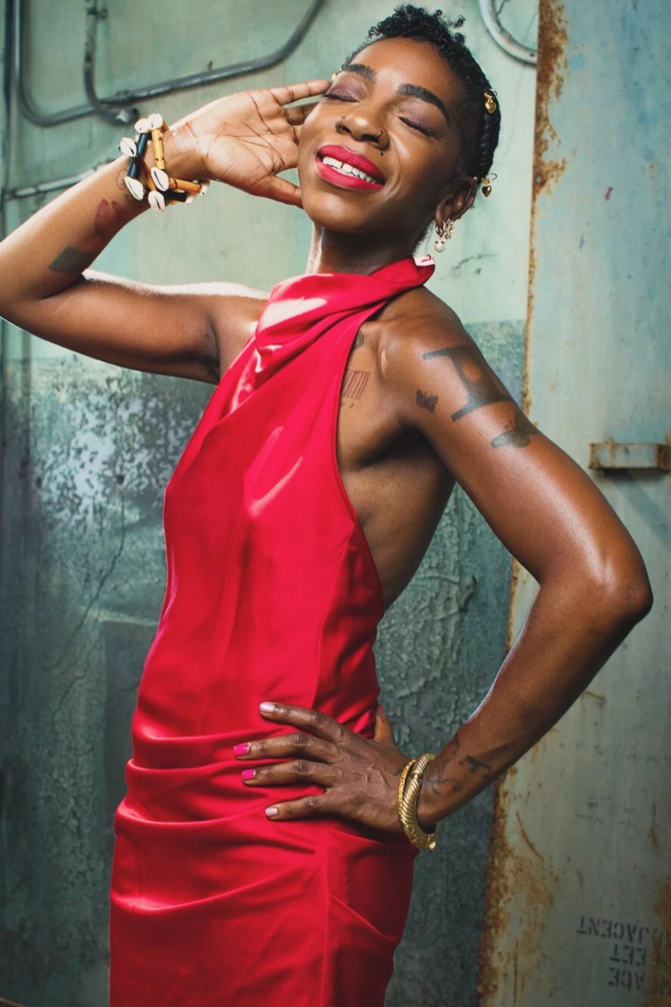 Cole Williams' styled with short curly hair, tattoos, and jewelry, wearing a sleeveless red satin dress, smiling with closed eyes and standing with one hand on her hip, in front of an industrial background.