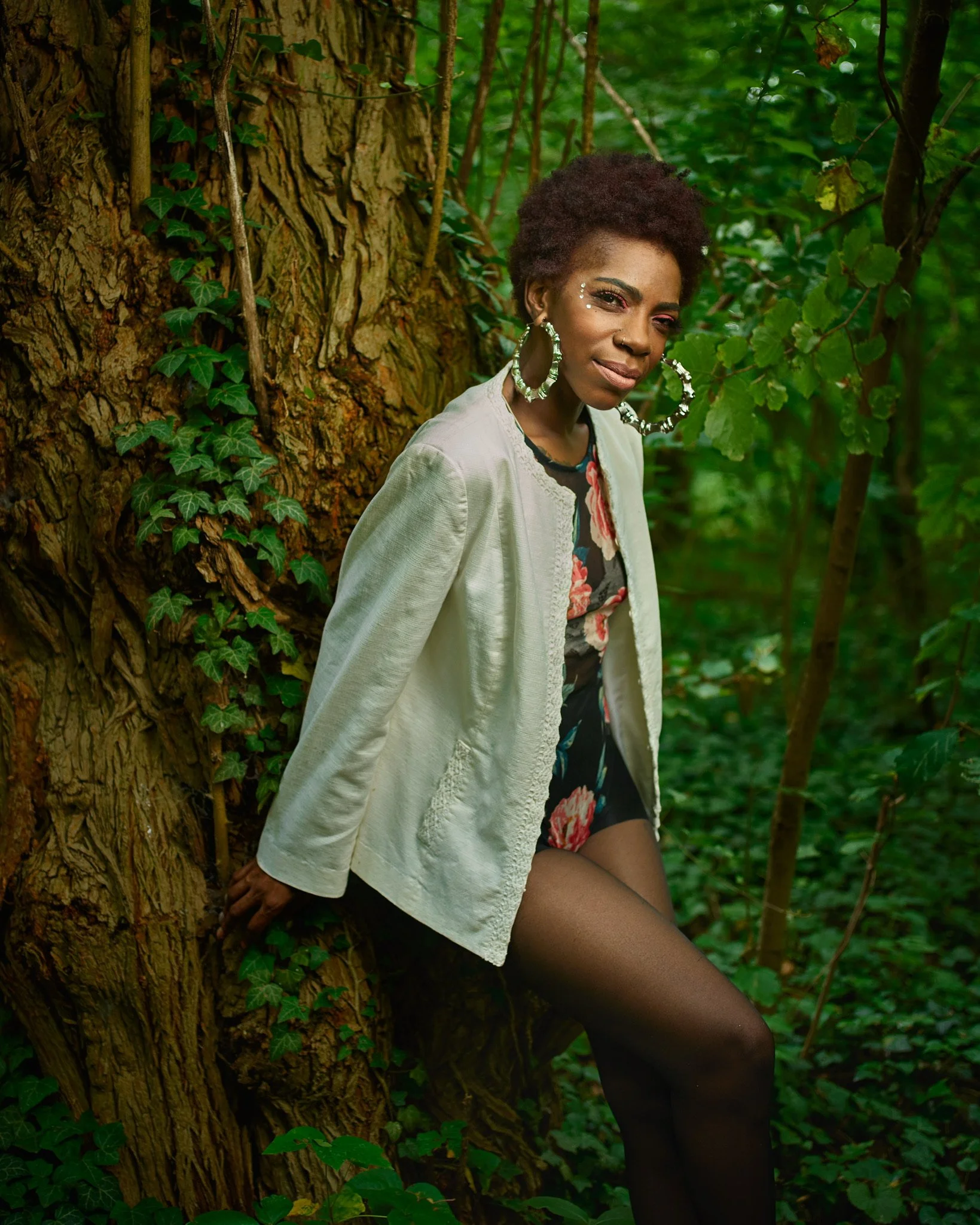A woman with short curly hair and large hoop earrings leaning against a tree trunk in a lush green forest, wearing a floral dress, a white jacket, and black tights.