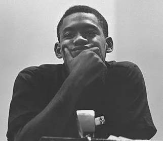 Curtis Muhammad as a young man with short hair, smiling and resting his chin on his hand, sitting at a table in a black and white photo.