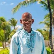 Anhk Ra Amenhotep, smiling, wearing a light blue embroidered shirt outdoors with palm trees and a clear blue sky in the background.