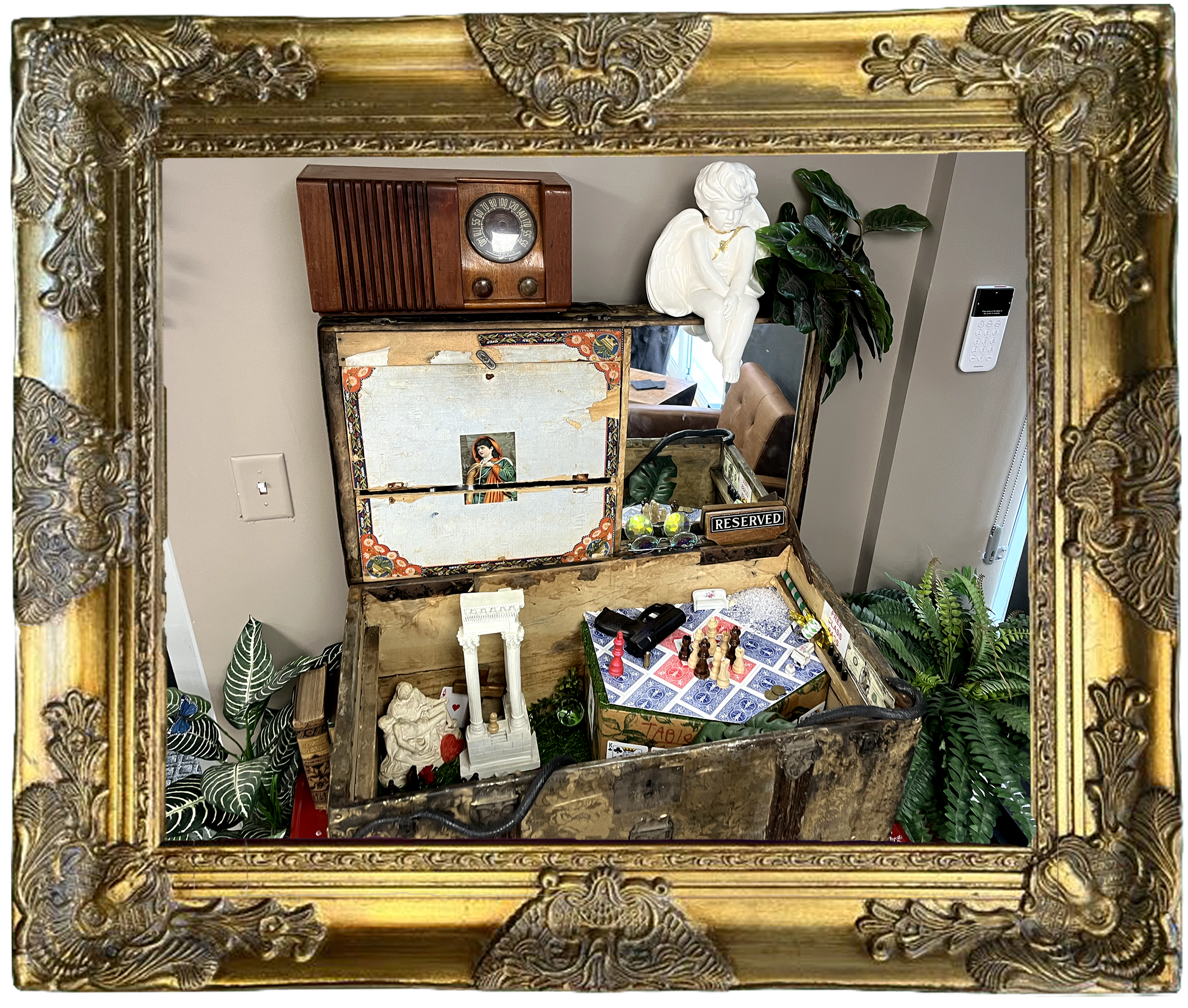 An ornate gold frame mirroring a cluttered antique trunk with vintage items, plants, and a figurine in a modern indoor setting.