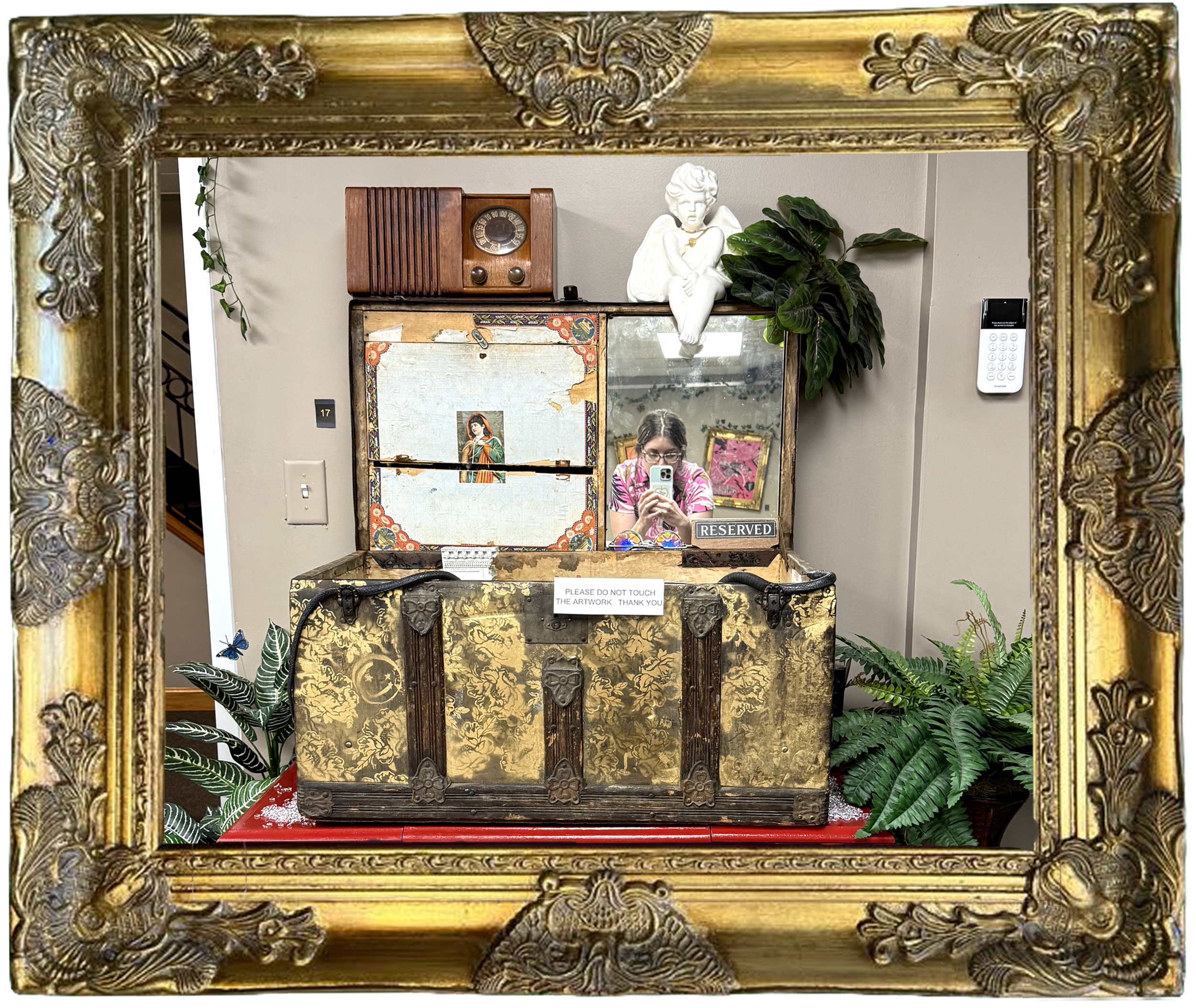 A person taking a photo of a decorative vintage box with a mirror, inside a gold ornate frame, surrounded by plants, a sculpture, and vintage items on top.