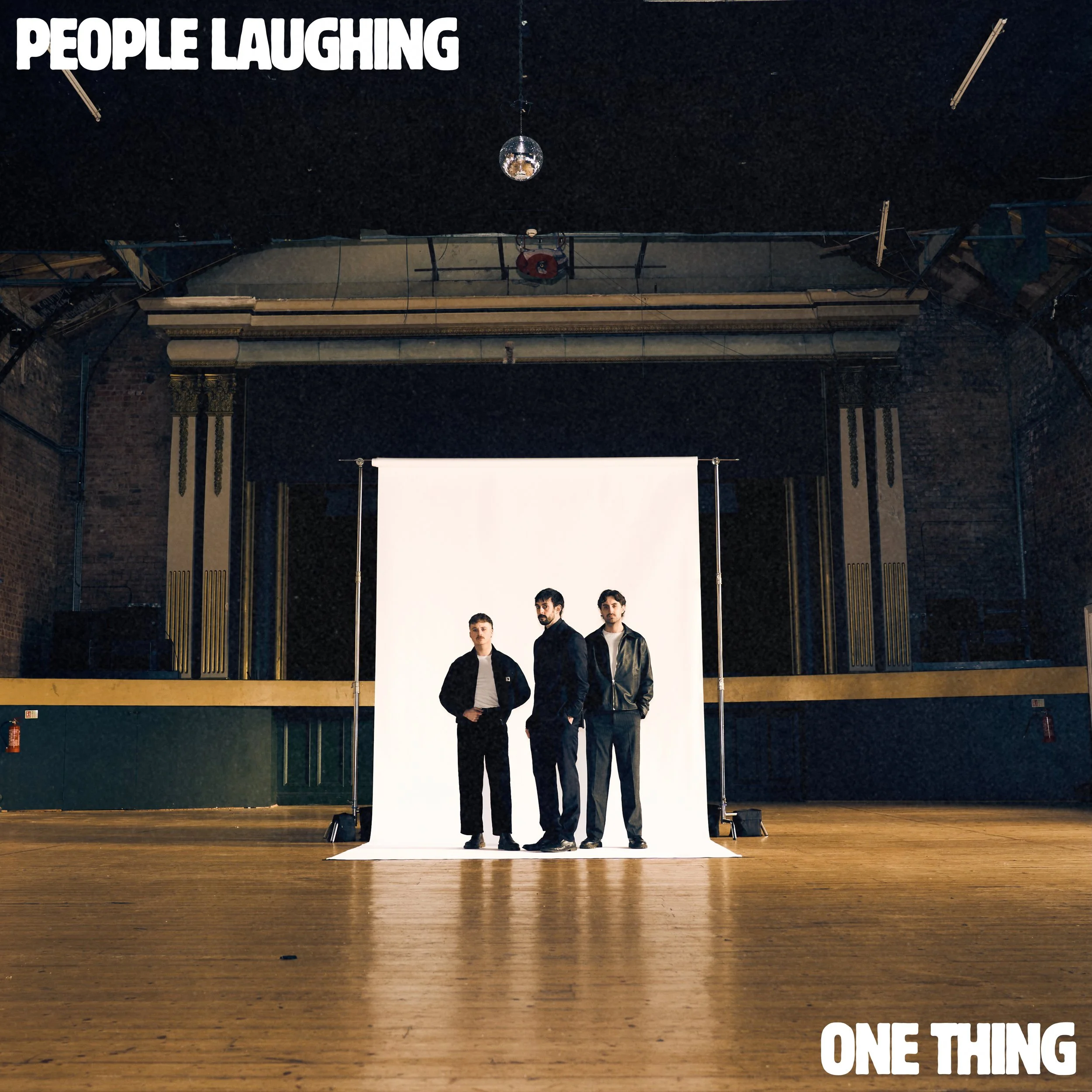 Three men standing in front of a white backdrop in a large empty room with a wooden floor, exposed brick walls, and high ceiling, with the words 'PEOPLE LAUGHING' in the top left corner and 'ONE THING' in the bottom right corner.