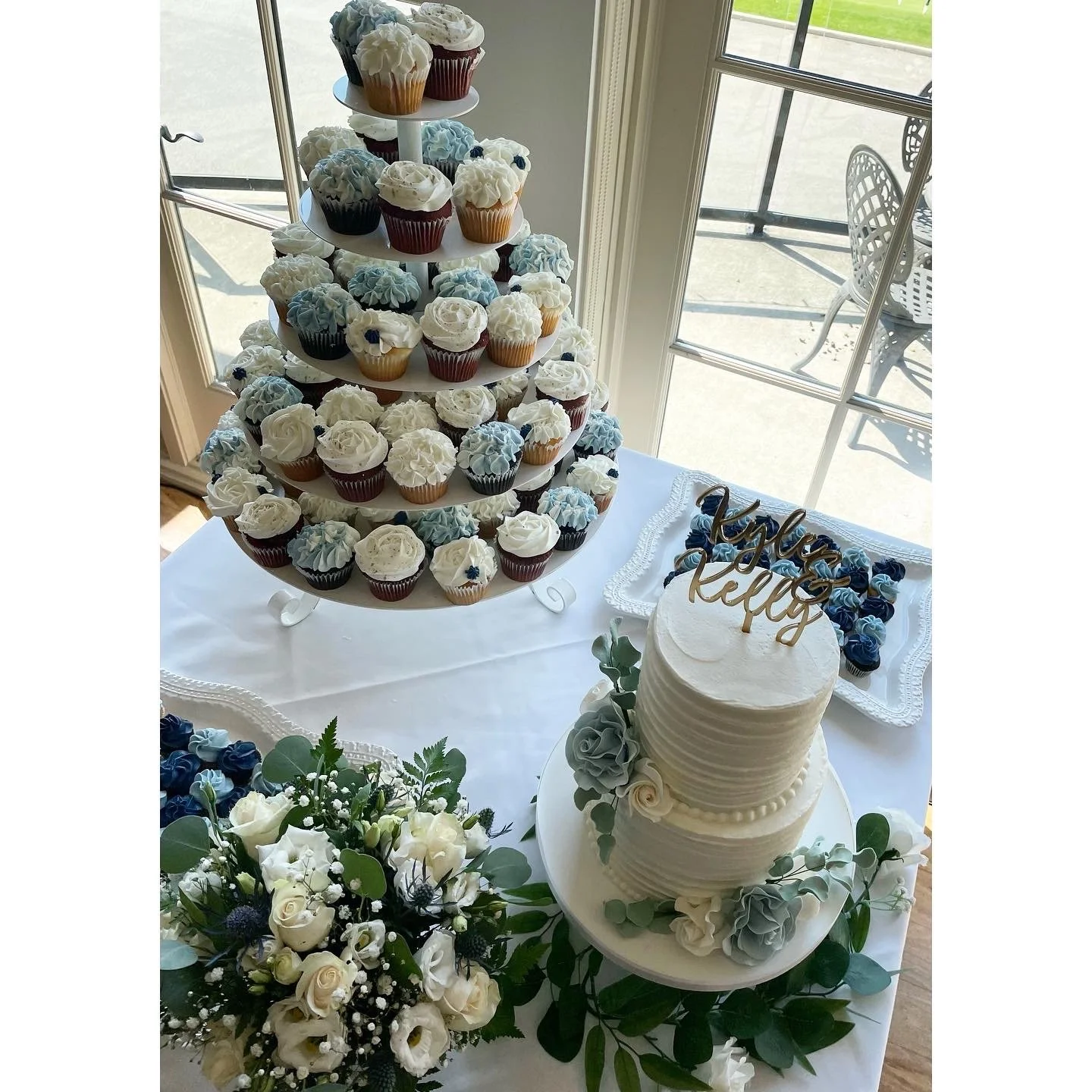 A wedding dessert table featuring a three-tiered cupcake stand with white and blue frosting cupcakes, a white wedding cake with floral decorations.