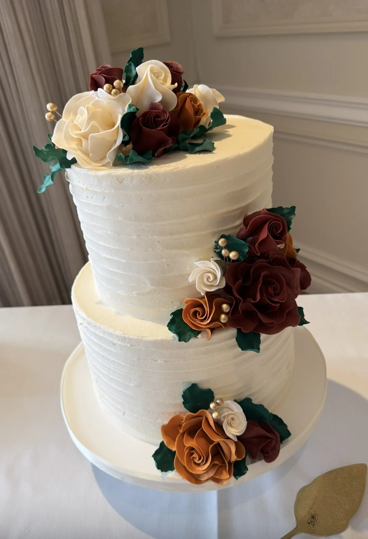 Two-tier white wedding cake decorated with colorful sugar roses, leaves, and gold pearl accents.