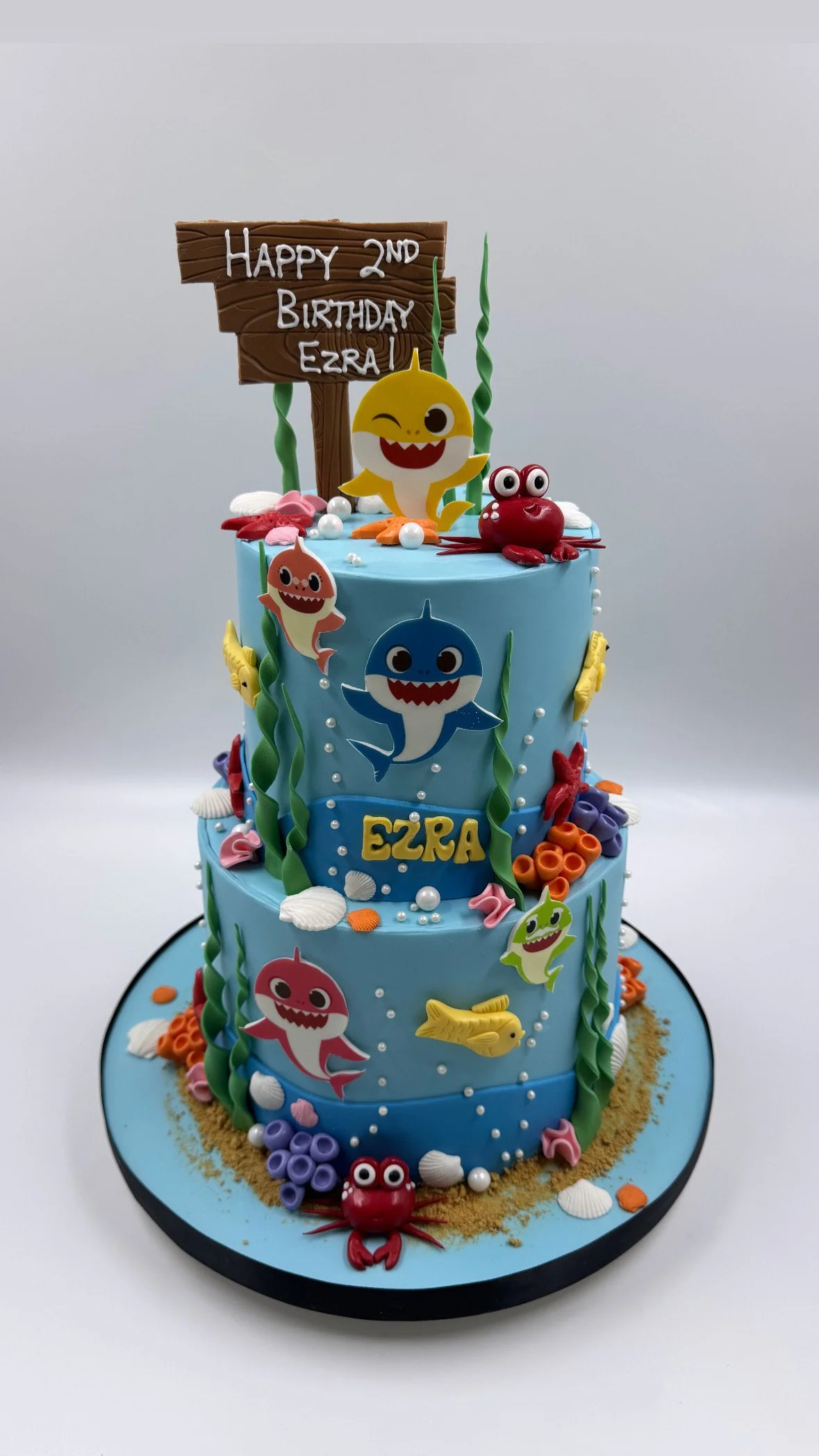 Three-tiered birthday cake with an underwater/ocean theme featuring cartoon sharks, fish, crabs, seashells, and seaweed decorations.