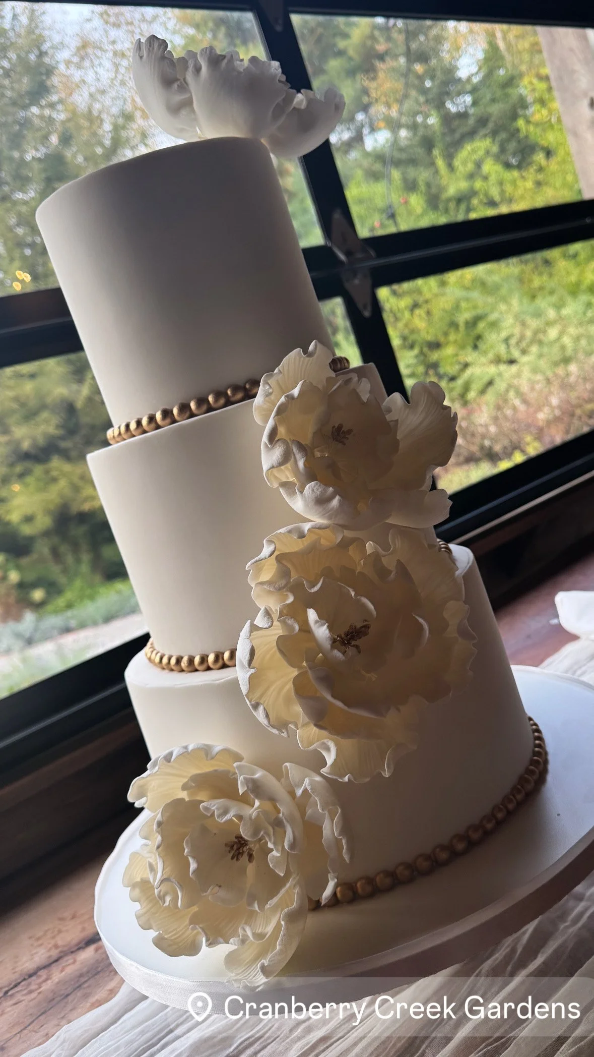 A three-tiered white wedding cake decorated with large white flowers and a gold beaded band around the middle tier, set on a white tablecloth with a view of greenery outside through a window.