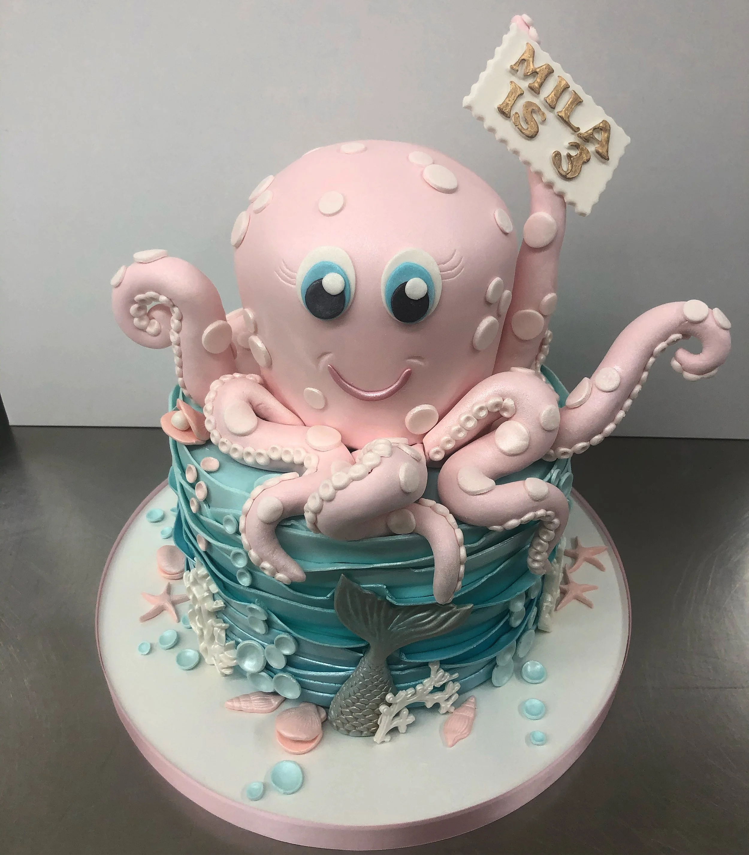 Decorative cake with an octopus theme, featuring a pink octopus with big blue eyes and a smiling face, holding a sign. The octopus is placed on a blue water-like base with a silver fish tail decoration and small shells and starfish around it.