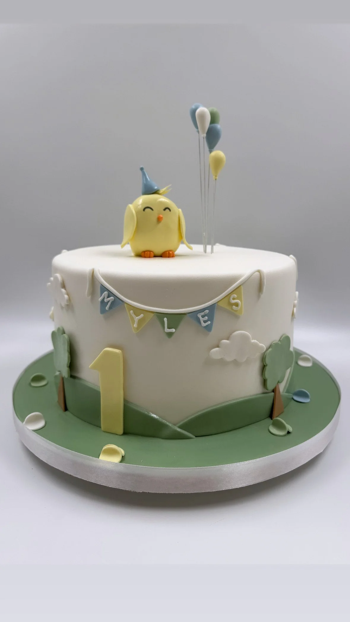 A birthday cake with a white top and green base, decorated with a banner, a yellow number one, a cute yellow chick figure wearing a blue party hat, a small cluster of balloons in pastel colors, and small fondant trees and clouds.