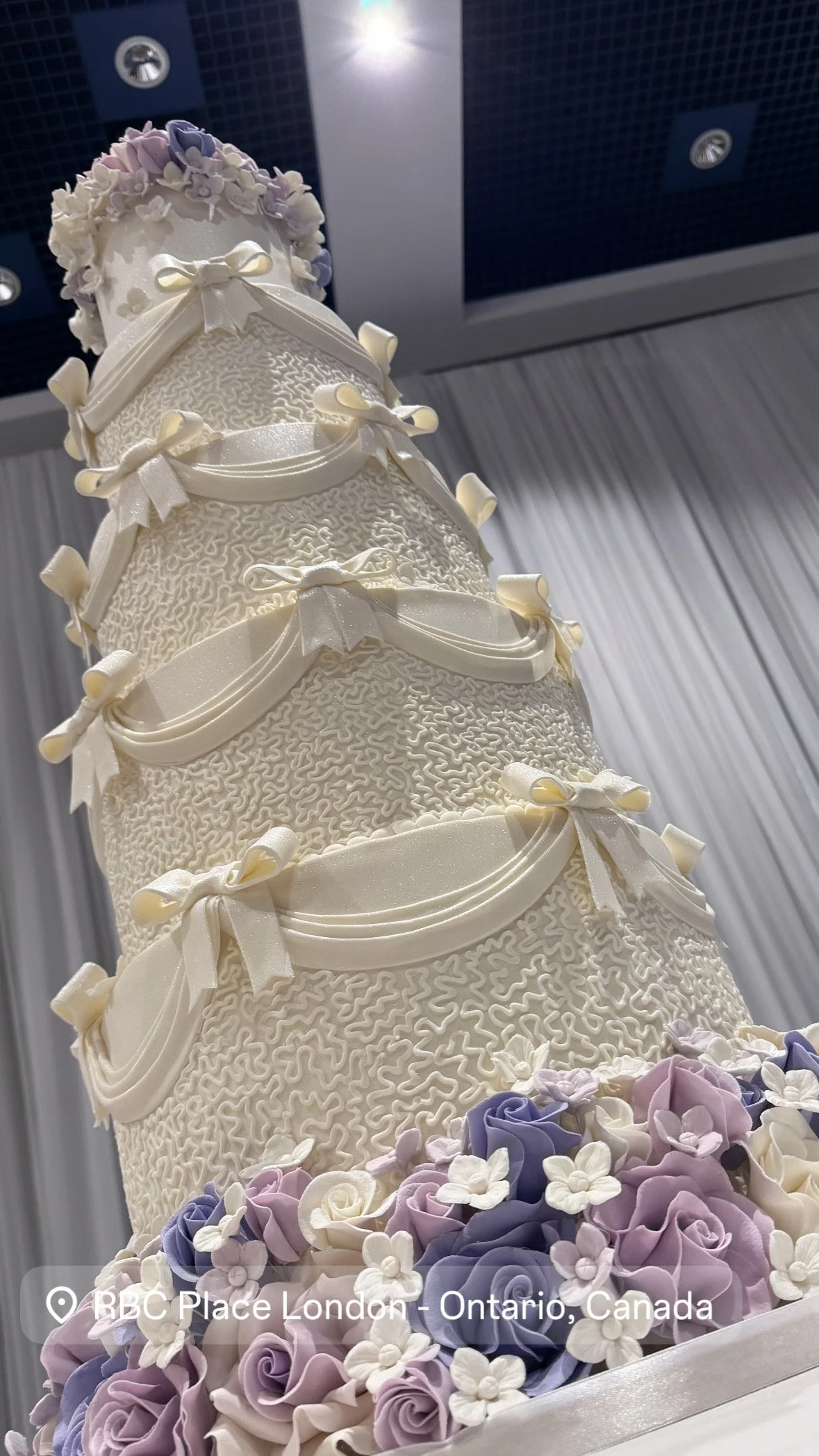 A tall, multi-tiered wedding cake decorated with white icing, bows, and floral accents in shades of purple, white, and cream, set against a background of white curtains and ceiling lights.