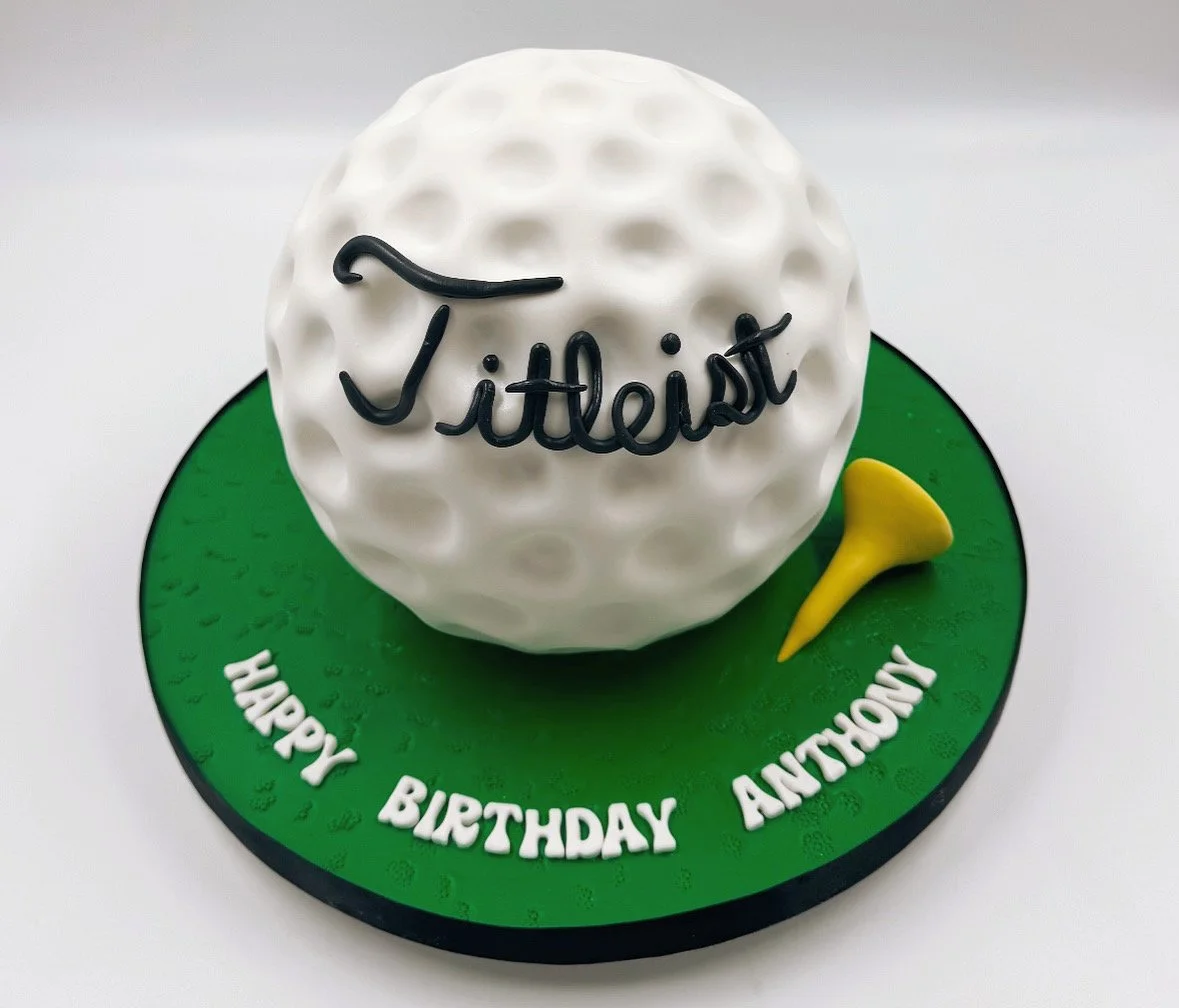 A golf-themed birthday cake with a white golf ball cake on top, a small yellow golf tee, and green icing on the base.
