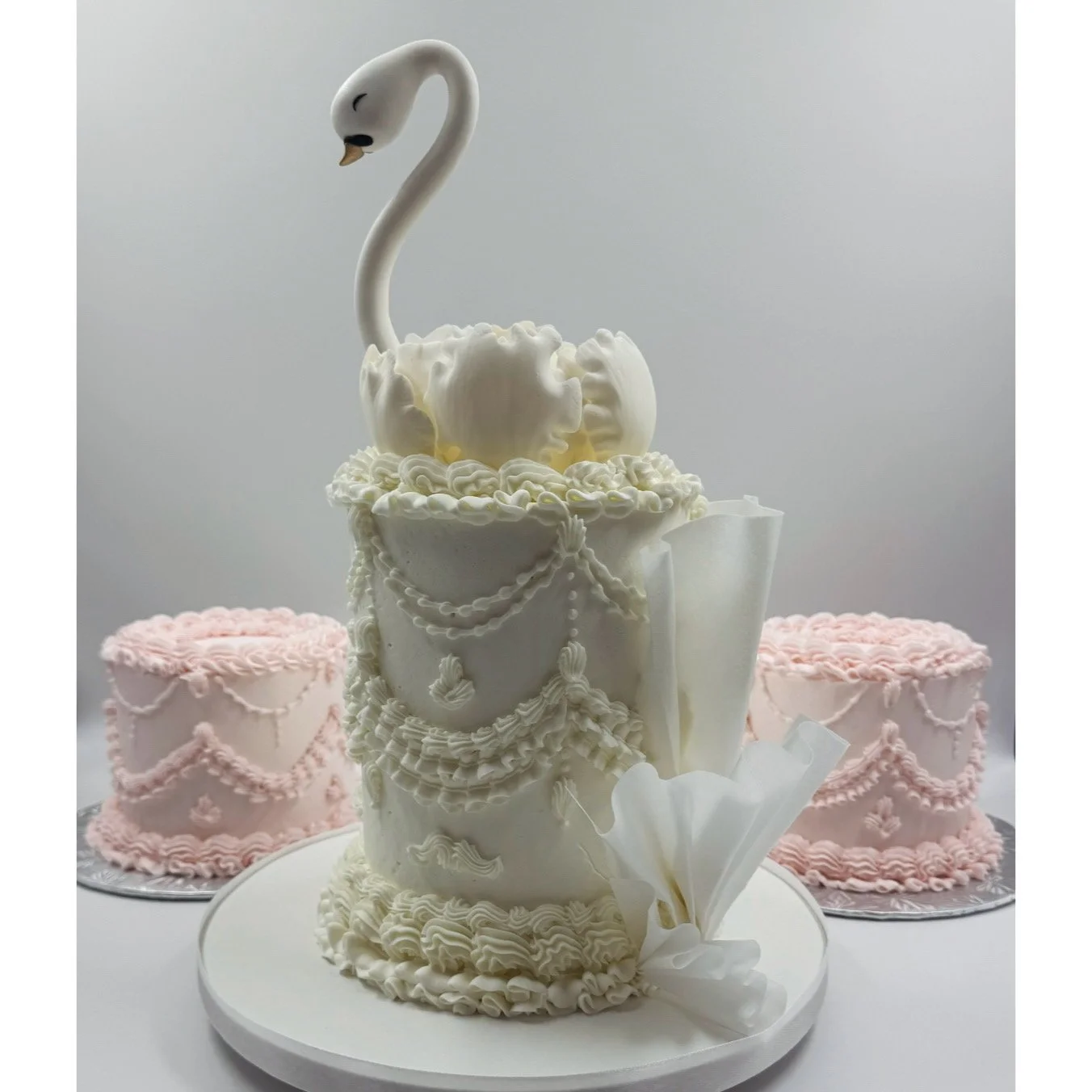 A white swan-shaped cake with detailed piping and decorations, surrounded by two pink decorated cakes.