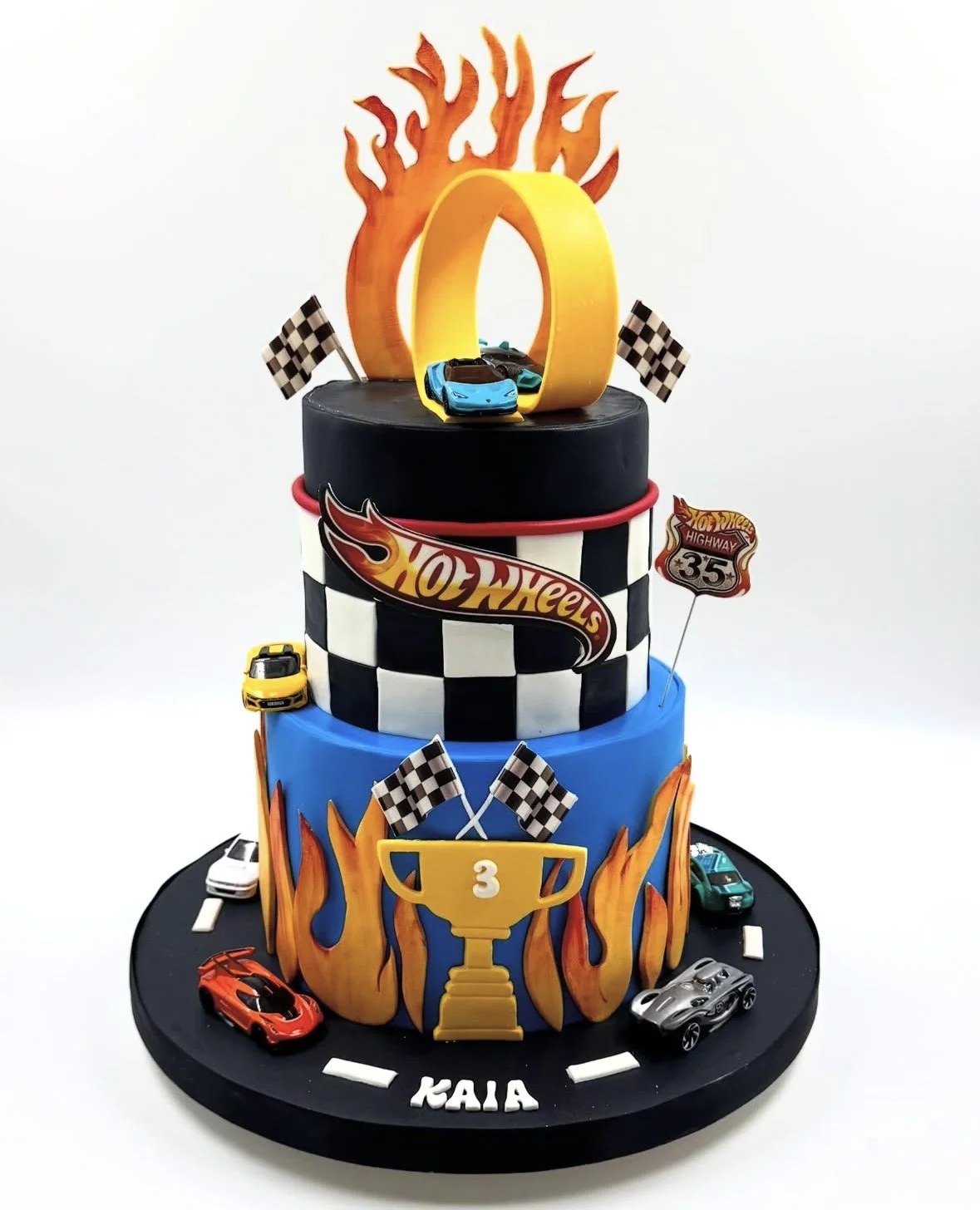 Three-tier race car themed birthday cake with Hot Wheels decorations, checkered flags, flames, a trophy with the number 3, and cars around the cake.