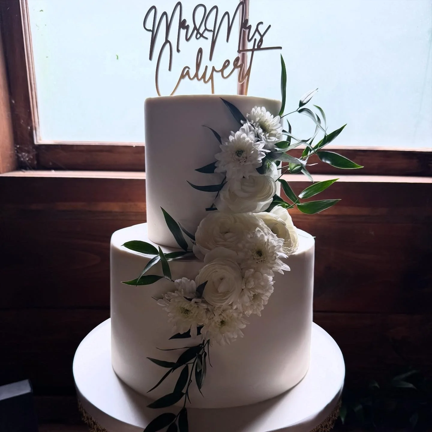 Two-tier white wedding cake decorated with white flowers and green leaves, cake topper on a white cake board.