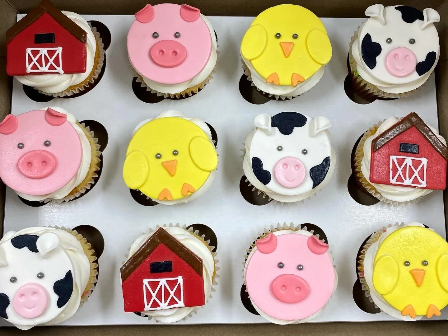 Decorative cupcakes with farm animal and barnhouse designs, including pigs, chicks, cows, and barn motifs, arranged in a box.