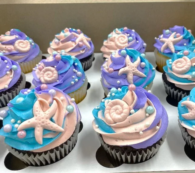 Cupcakes decorated with ocean-themed pastel-colored frosting, starfish, shells, and pearl-like decorations.