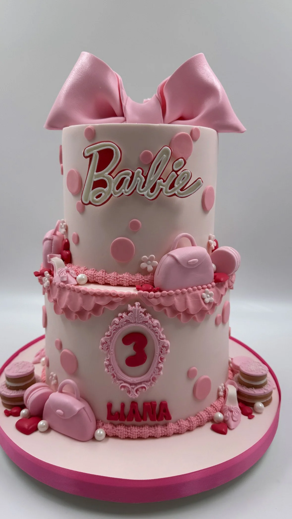 A two-tier pink and white birthday cake decorated with fondant and edible embellishments. The top tier features a pink bow and the name "Barbie", with pink polka dots. The bottom tier has a pink frame  with pink and white accessories like handbags, h