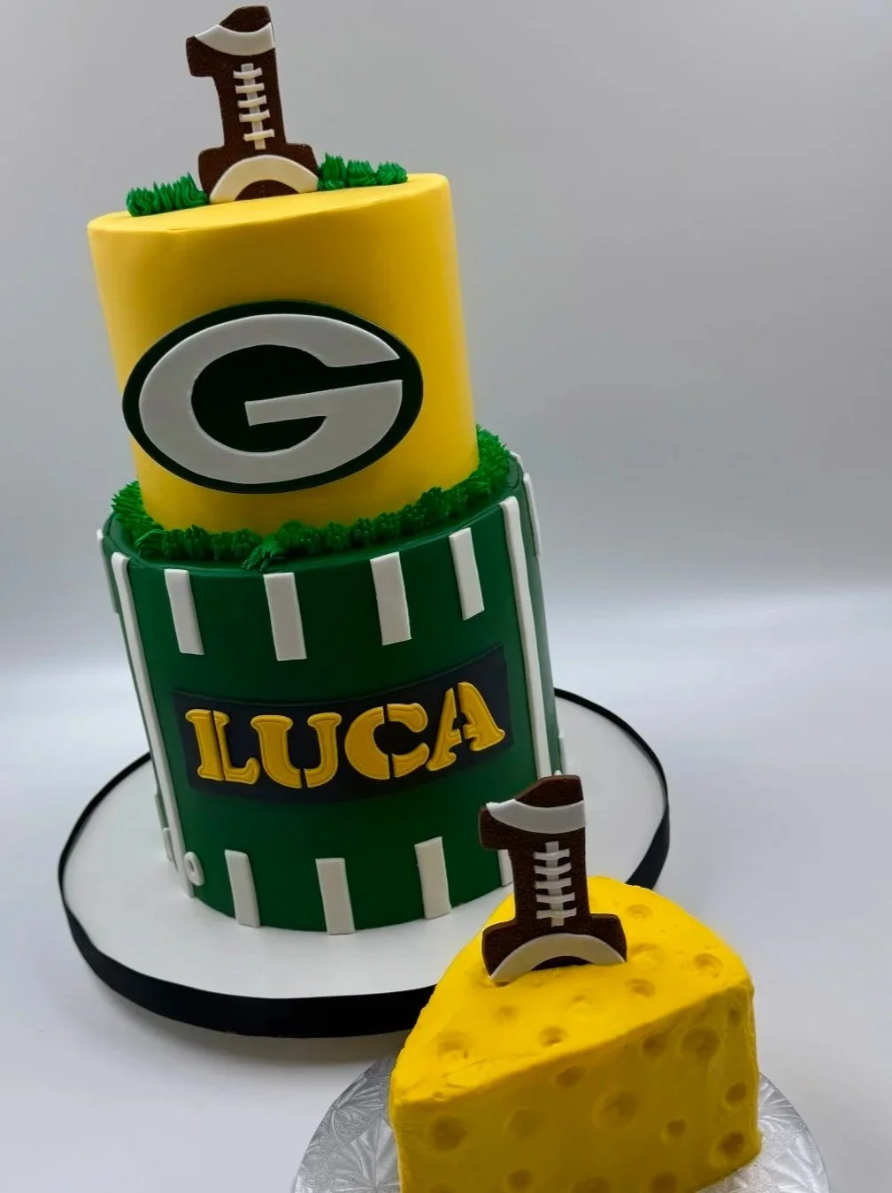 A two-tiered cake with Green and yellow football-themed decorations, featuring the letter G. The cake is topped with football-shaped decorations and a football field design.