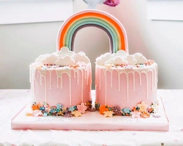Two pink cakes with white drip icing and colorful floral decorations, topped with a rainbow made of fondant, on a white cake board.