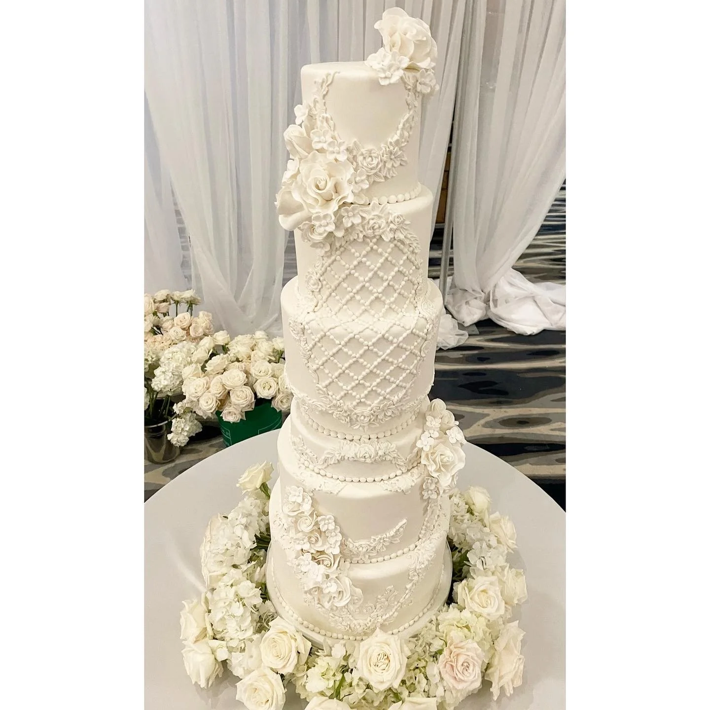 Five-tier white wedding cake decorated with intricate floral and lattice patterns, topped with sugar flowers, surrounded by white roses and hydrangeas.