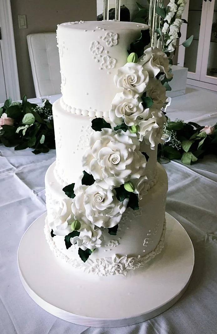 A tall, three-tiered white wedding cake decorated with cascading white roses, green leaves, and intricate white piping designs.