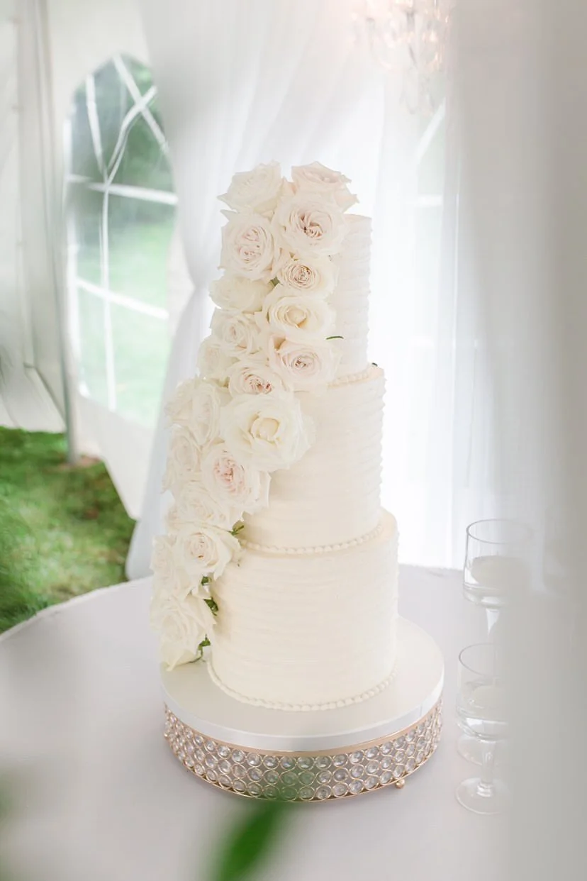A tall, elegant white wedding cake decorated with cascading white roses, placed on a round table with a decorative rhinestone border.