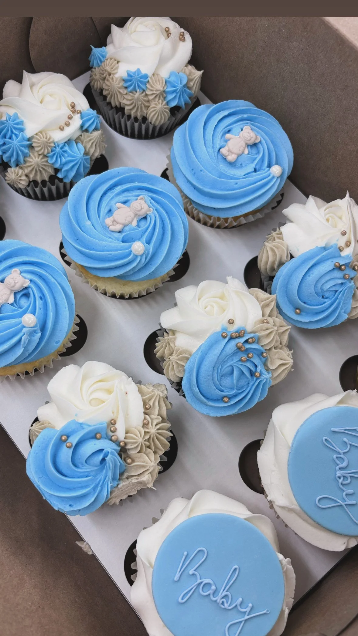 A tray of cupcakes decorated with blue, white, and beige frosting, some featuring small teddy bear decorations and pearl-like sprinkles, with a blue fondant topper that says 'Baby'.