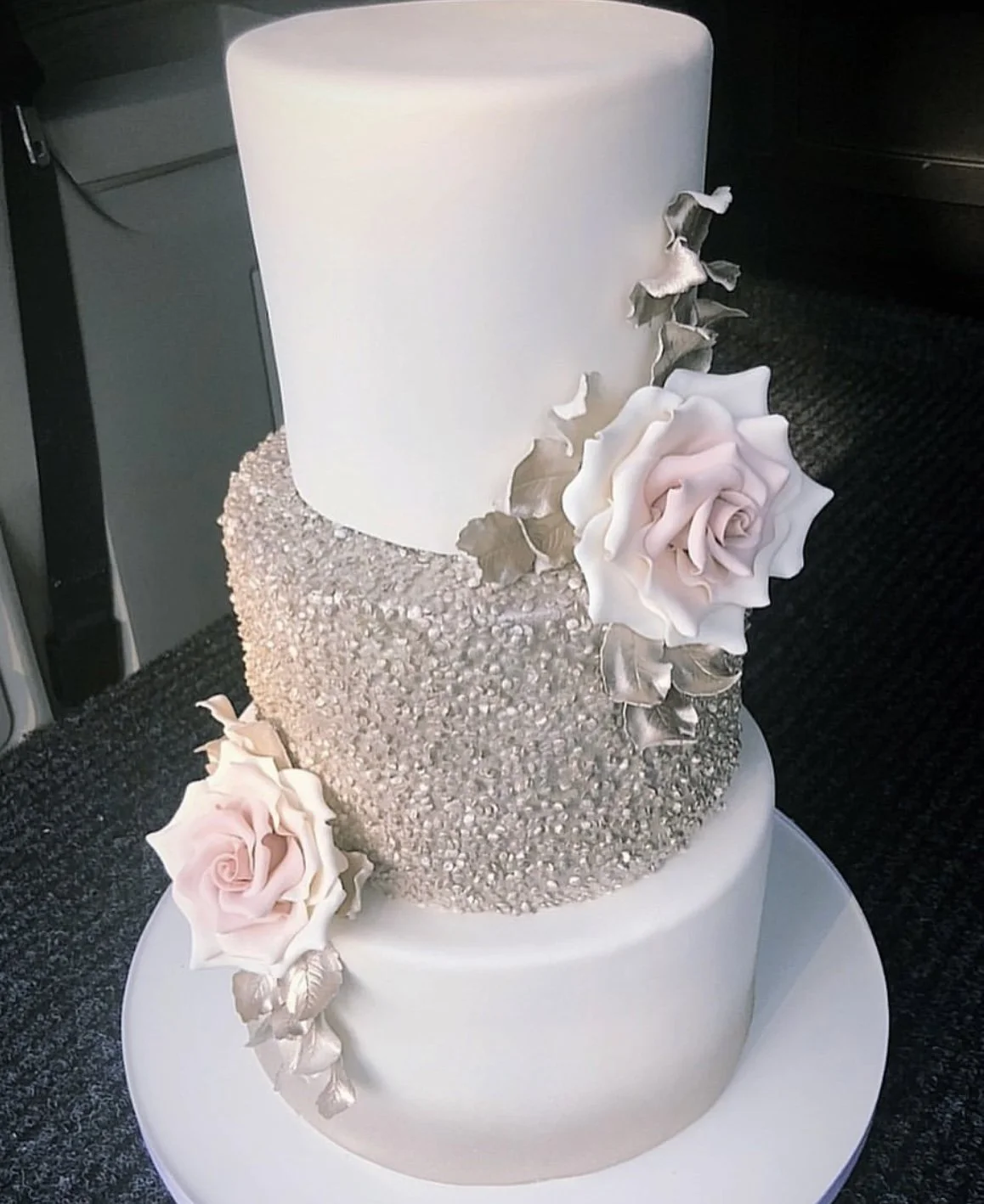 A three-tiered white wedding cake with textured silver accents and pink roses, decorated with silver leaves.