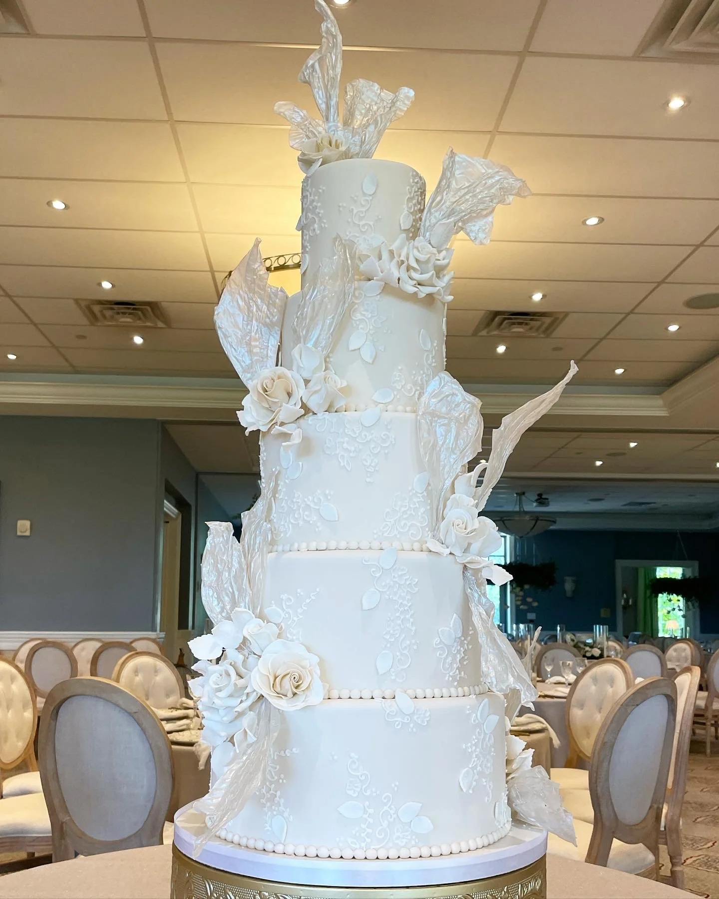 A tall, elegant white wedding cake with five tiers, decorated with white fondant flowers, leaves, and intricate patterns, topped with white floral decorations.